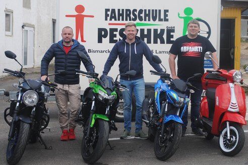Drei Fahrlehrer mit Motorrädern und Roller vor dem Fahrschulanhänger